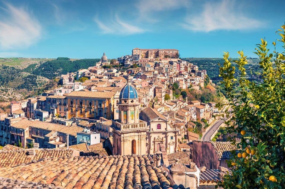 Una vista panorámica de un pueblo histórico en la ladera con tejados de terracota y una iglesia de cúpula azul bajo un cielo azul brillante.