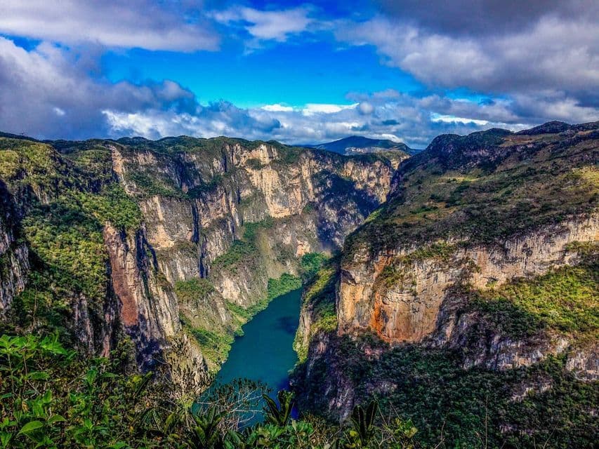 Un fiume turchese scorre attraverso un canyon profondo con ripide scogliere ricoperte di vegetazione sotto un cielo blu con nuvole sparse.