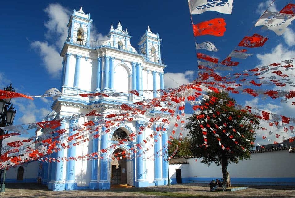 Una chiesa bianca e blu è decorata con festoni di bandierine di carta rosse e bianche in una piazza sotto un cielo azzurro limpido.