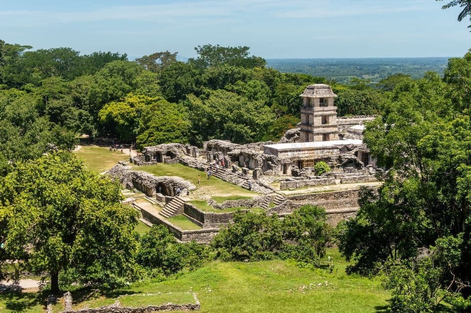 Una vista de gran angular de antiguas ruinas de piedra, incluyendo una torre prominente, rodeada por una densa jungla verde bajo un cielo despejado.