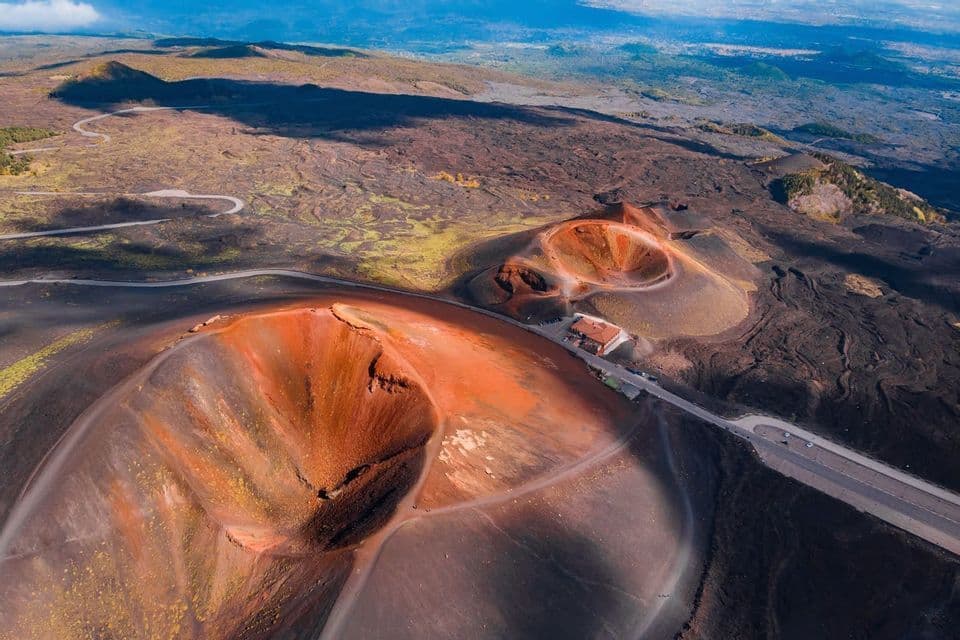 An aerial view of reddish volcanic craters and a winding road cutting through a dark, rocky landscape under a blue sky.