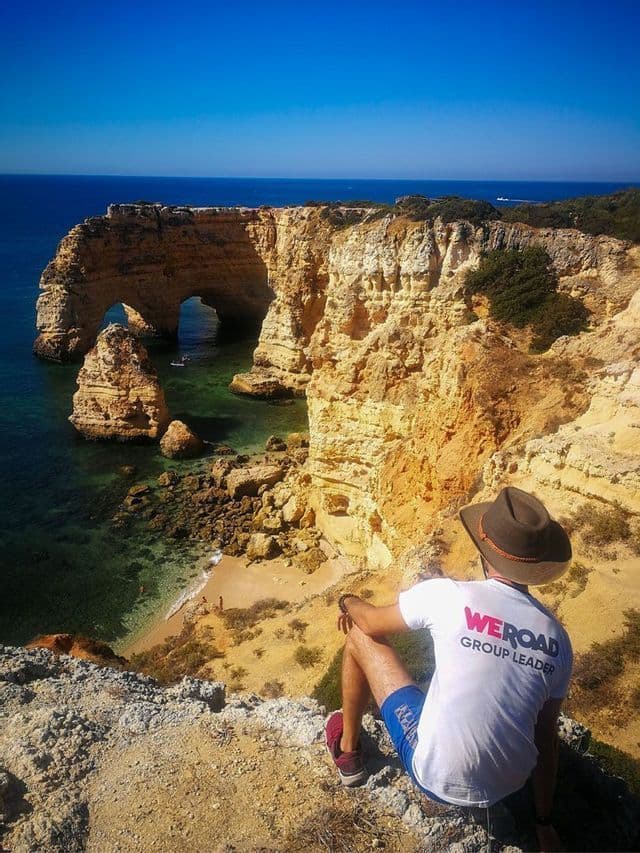 Ein WeRoad-Gruppenleiter mit Hut sitzt auf einer Klippe mit Blick auf eine felsige Küste mit natürlichen Felsbögen und einem kleinen Strand.