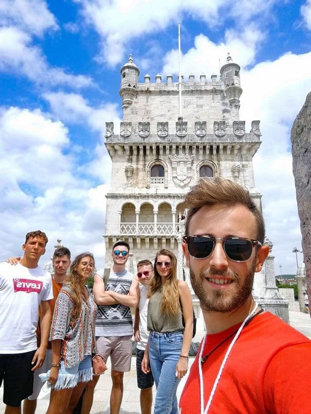 Un homme en lunettes de soleil prend un selfie avec un groupe WeRoad lors d'un voyage devant une tour en pierre historique sous un ciel bleu nuageux.