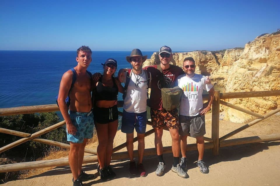 Un gruppo WeRoad di cinque persone in posa per una foto su un punto panoramico costiero soleggiato, con l'oceano blu e le scogliere sullo sfondo.
