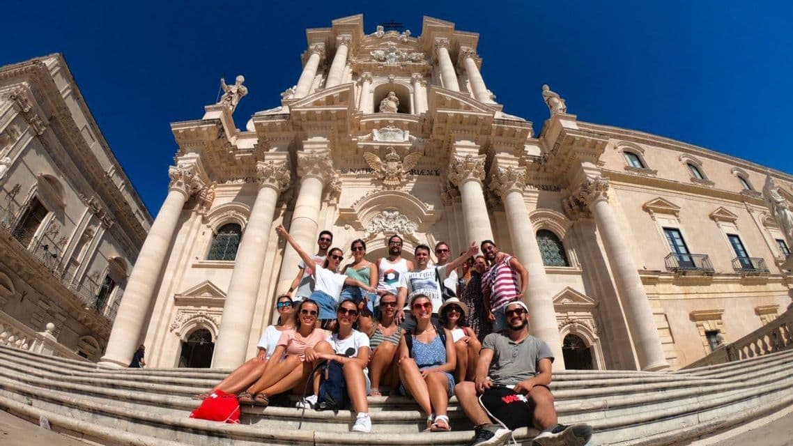 Un grupo de viaje de WeRoad posando para una foto en las escalinatas de un edificio barroco ornamentado bajo un cielo azul despejado.