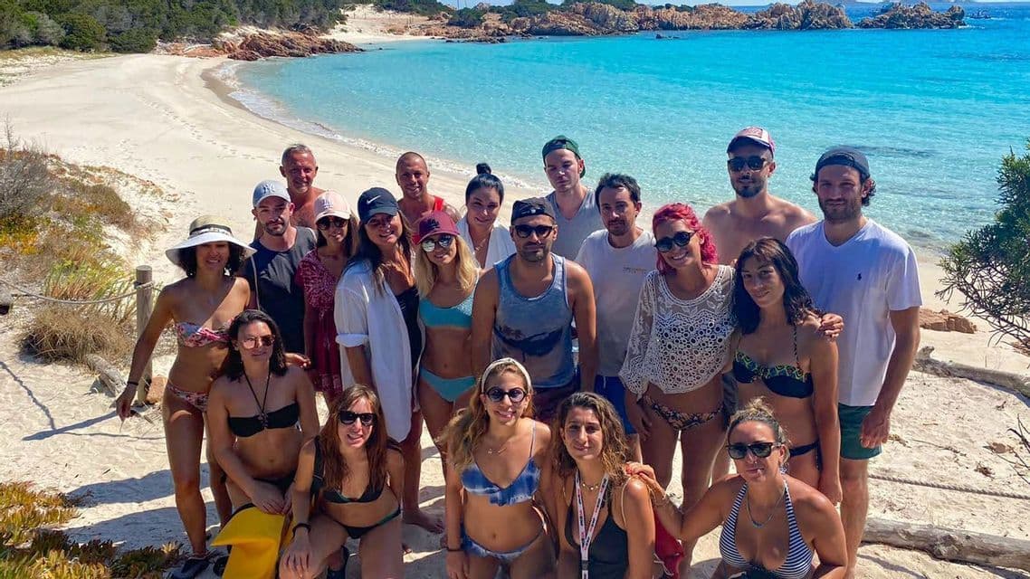 Un gruppo WeRoad in posa per una foto su una spiaggia di sabbia bianca con acqua turchese cristallina e una costa rocciosa sullo sfondo
