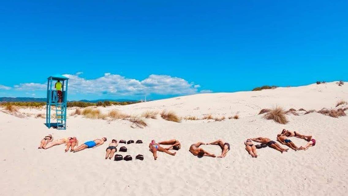 Un grupo de viaje de WeRoad deletrea la palabra 'WEROAD' con sus cuerpos, tendidos en una playa de arena blanca bajo un cielo azul claro.