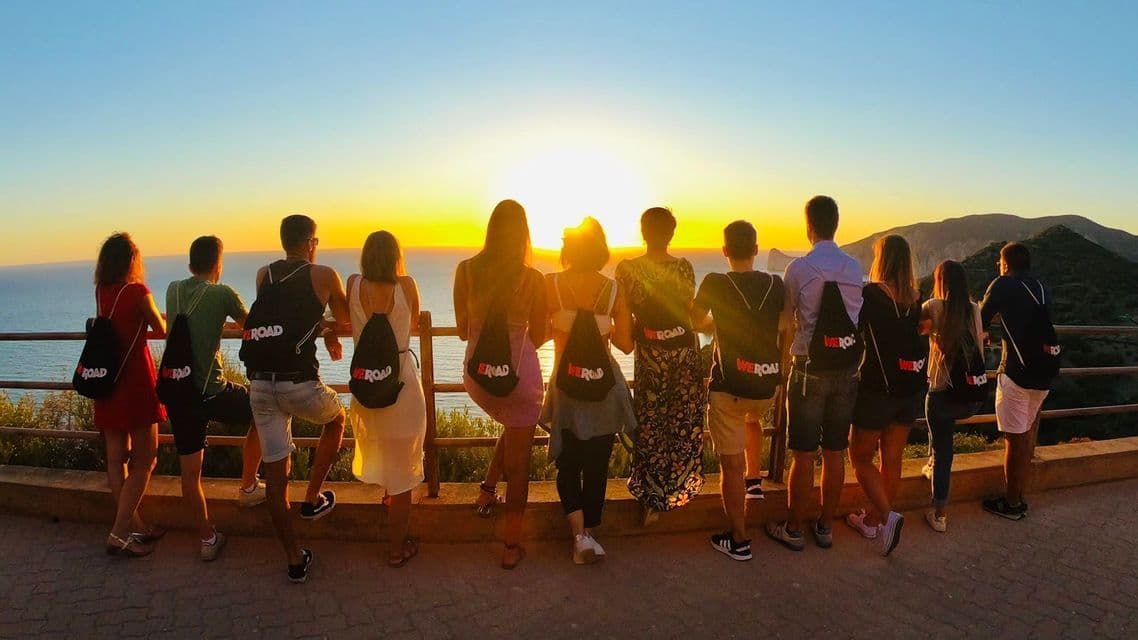 Un grupo de WeRoad, visto de espaldas, contempla el atardecer sobre el mar desde un mirador panorámico.