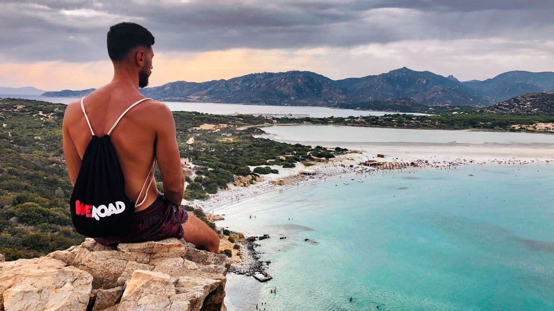 Un hombre con una mochila WeRoad se sienta en un acantilado rocoso, mirando una playa concurrida con agua turquesa y montañas.
