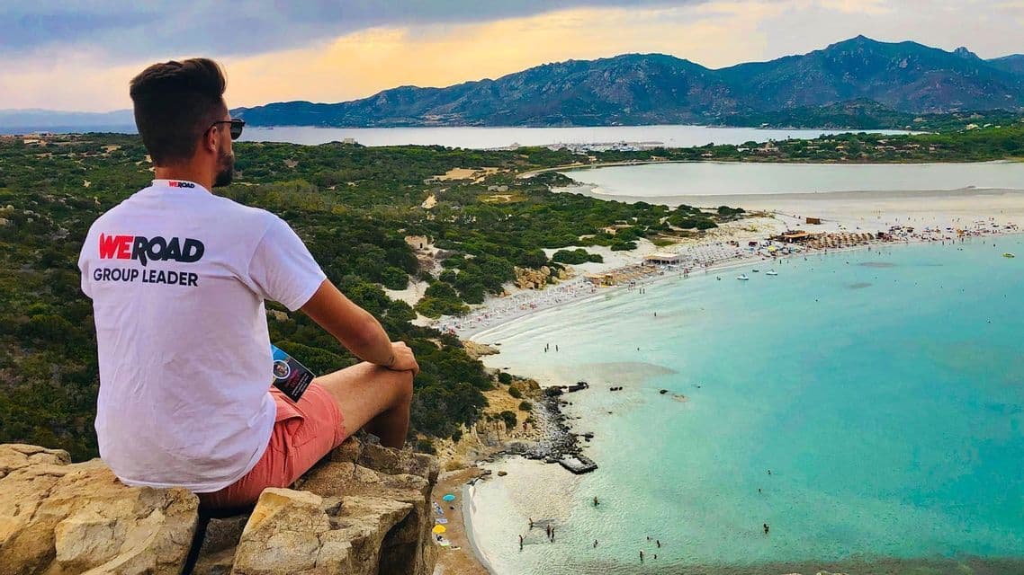 Un capogruppo WeRoad siede su una scogliera rocciosa che domina una baia con una spiaggia di sabbia bianca e acqua turchese.