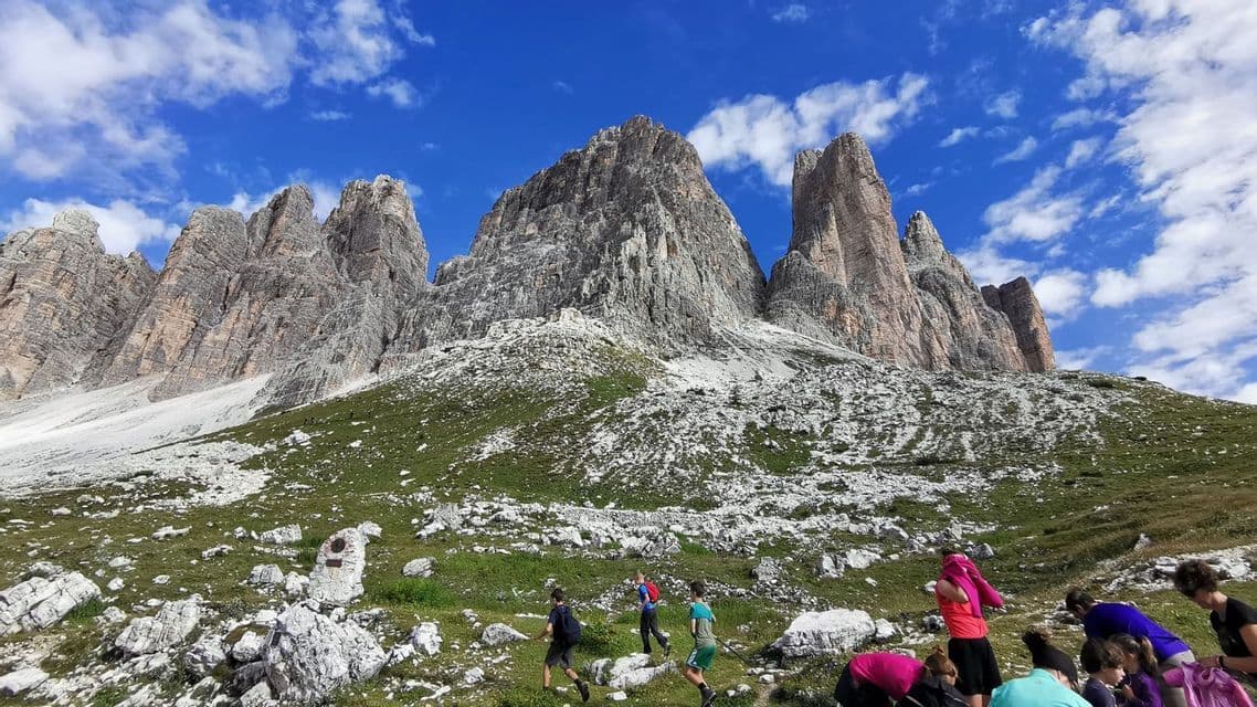 Eine WeRoad-Gruppenreise beim Wandern an einem grünen, felsigen Berghang mit zerklüfteten Gipfeln vor blauem Himmel mit weißen Wolken.
