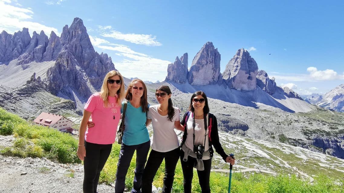 A WeRoad group trip of four women smiling on a mountain trail, with a jagged, rocky mountain range in the background.