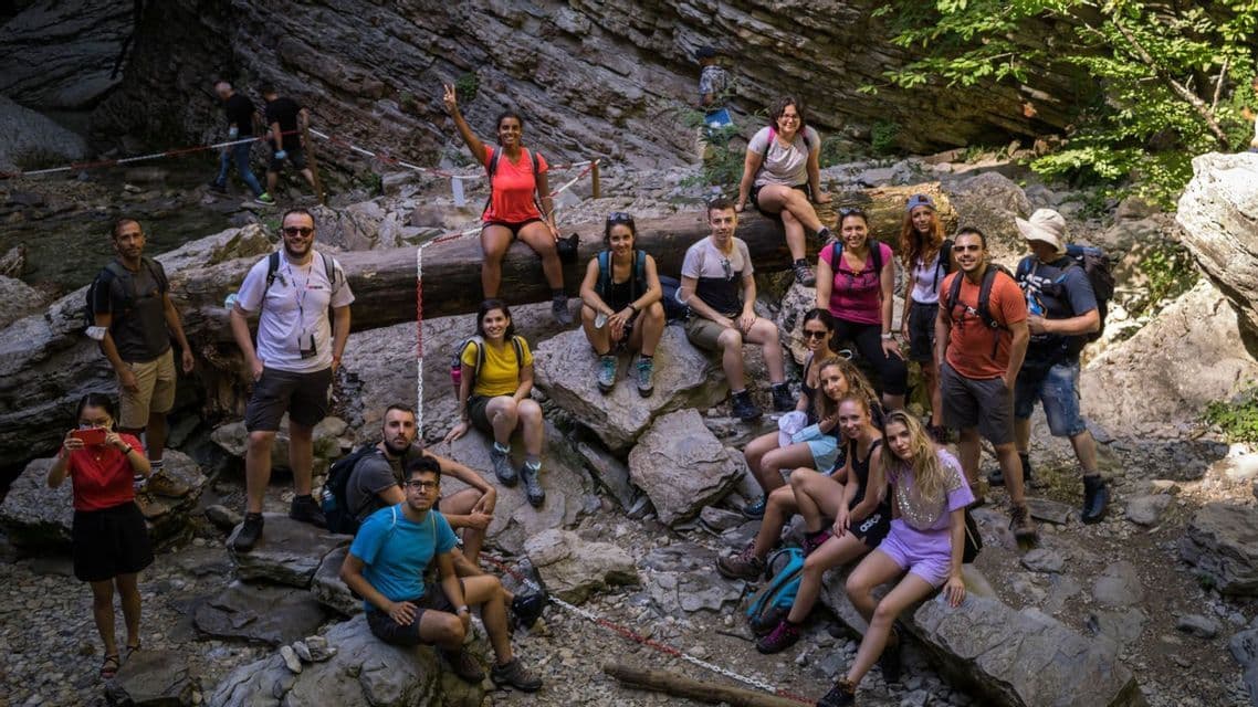A large WeRoad group trip poses for a photo, sitting and standing on rocks and a fallen log in a canyon.