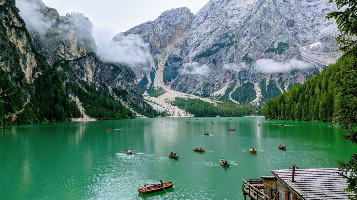 Menschen in hölzernen Ruderbooten paddeln über einen türkisfarbenen Alpensee, mit bewaldeten Hängen und felsigen Bergen im Hintergrund.