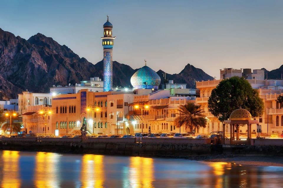 An illuminated mosque and city skyline on a waterfront at dusk, with rugged mountains rising behind the buildings.