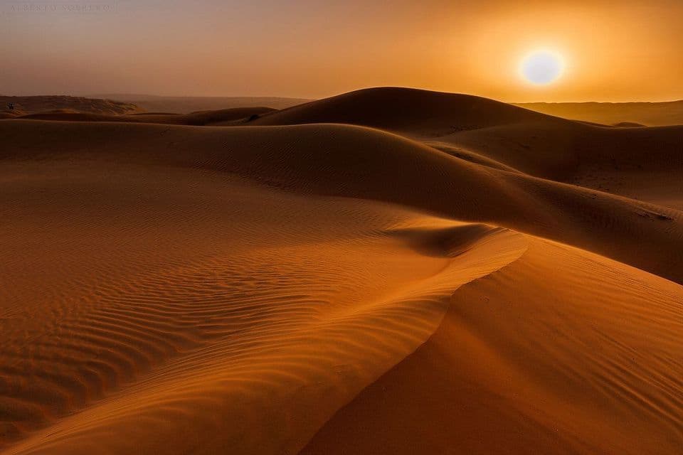Vast, rolling sand dunes with wind-swept ripples are illuminated by a setting sun in an orange sky.