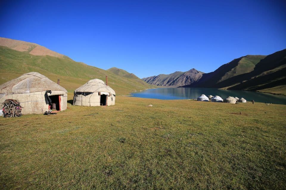 Un campo di yurte è allestito su una pianura erbosa accanto a un lago tranquillo, con dolci montagne verdi e marroni sotto un cielo azzurro chiaro.