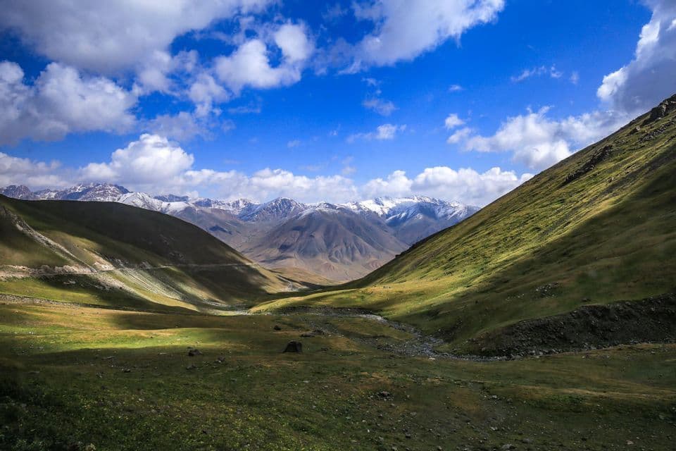 Una vasta valle verde si trova tra due grandi colline, con una lontana catena di montagne innevate sotto un cielo azzurro e nuvole bianche e soffici.