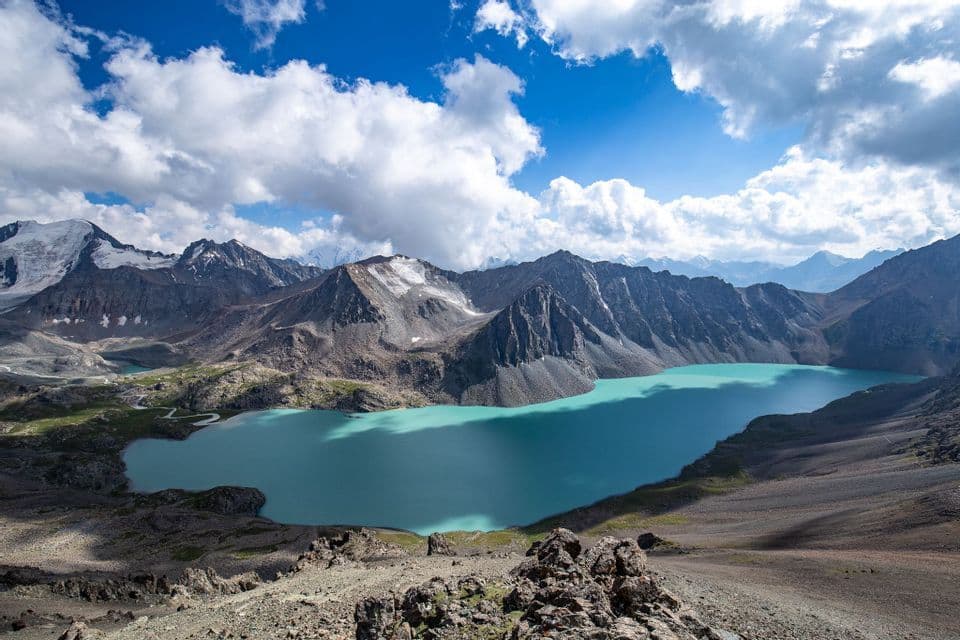 Un vivace lago alpino turchese circondato da montagne rocciose e innevate sotto un cielo azzurro parzialmente nuvoloso.