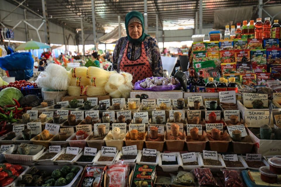 Eine Frau mit Kopftuch und Schürze steht hinter einem geschäftigen Marktstand, der Gewürze, Nudeln und andere Lebensmittel verkauft.