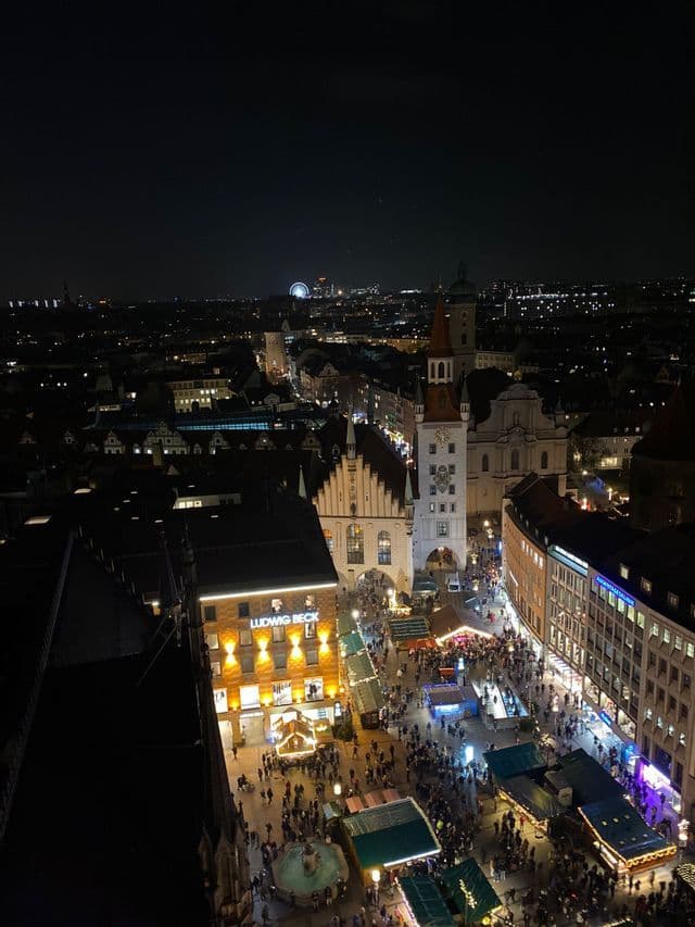 Una vista aerea di una vivace piazza cittadina di notte, piena di persone che camminano tra bancarelle del mercato ed edifici storici illuminati.