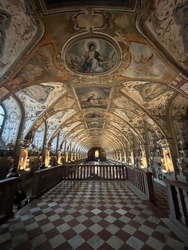 L'interno di una lunga sala con un soffitto a volta affrescato e ornato, sculture illuminate e un pavimento a scacchiera.