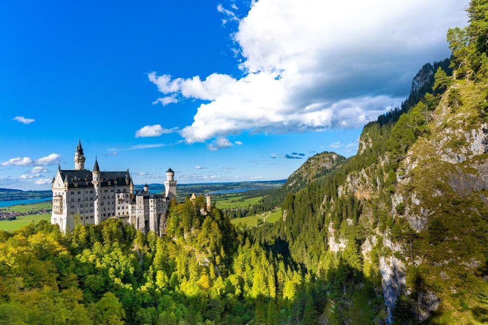 Un grande castello con torri si erge su una collina boscosa, dominando una vasta valle verde e montagne sotto un cielo azzurro con nuvole bianche.