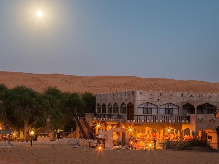 Au crépuscule, sous une pleine lune, un lodge désertique illuminé et arboré est niché contre des dunes de sable.