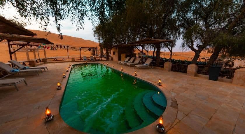 Une piscine aux eaux vertes, éclairée par de petites lanternes, est située dans une oasis désertique avec des chaises longues et des dunes de sable en arrière-plan.