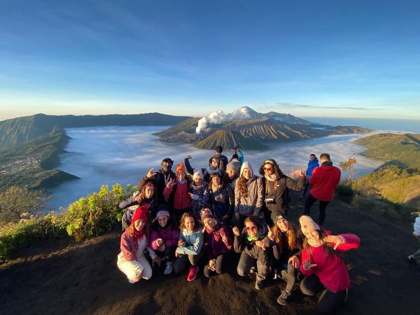 A WeRoad group trip poses on a mountain viewpoint at sunrise, overlooking smoking volcanoes and a sea of clouds.