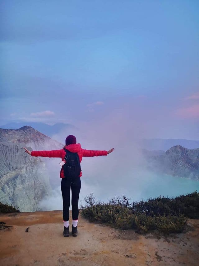A person wearing a red jacket and backpack stands on a cliff's edge with arms outstretched, overlooking a misty volcanic crater lake at dawn.
