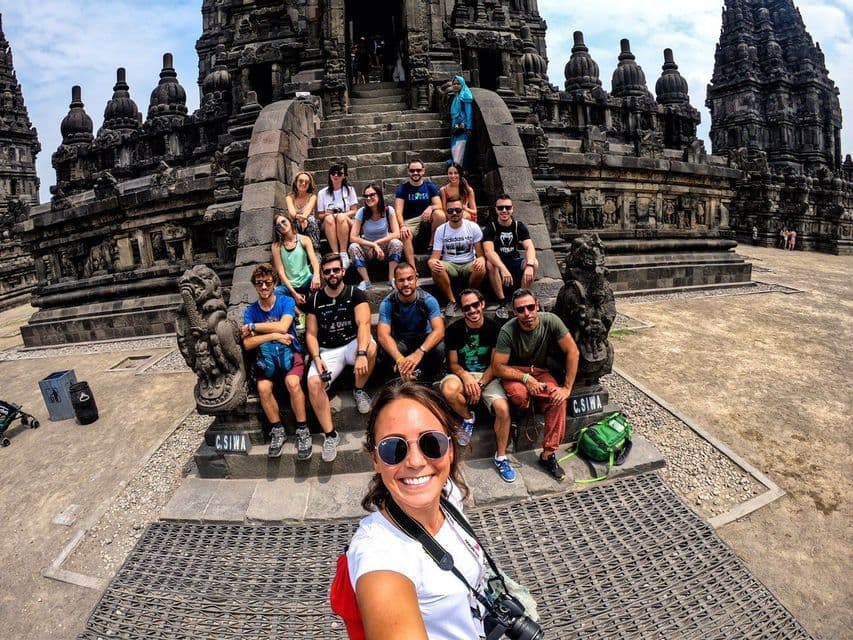 A woman takes a selfie with her WeRoad group trip, who are sitting together on the stone steps of an ornate temple.