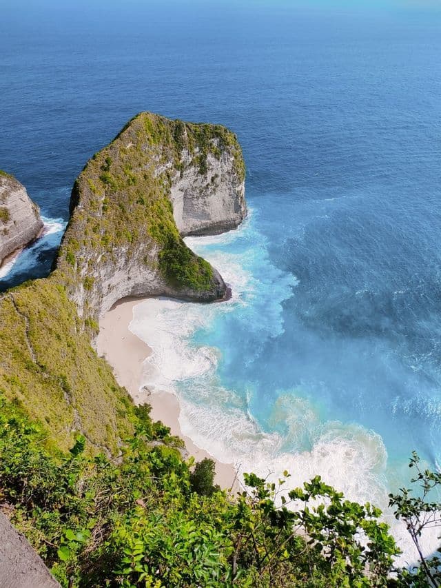 A high-angle view of a green-topped cliff formation over a secluded white sand beach with turquoise waves crashing on the shore.