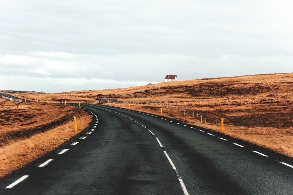 A paved road curves through a rural landscape of orange, dry grass, with a lone house on a hill in the distance.