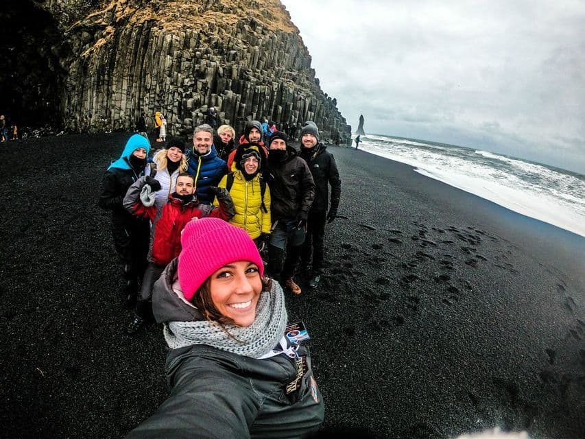 Una mujer con un gorro rosa se toma una selfie con un viaje en grupo de WeRoad en una playa de arena negra junto a formaciones rocosas de basalto.