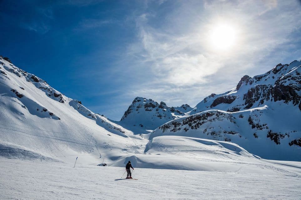 Un esquiador solitario se detiene en una vasta ladera nevada en un valle de montaña, bajo un sol radiante y un cielo azul claro.