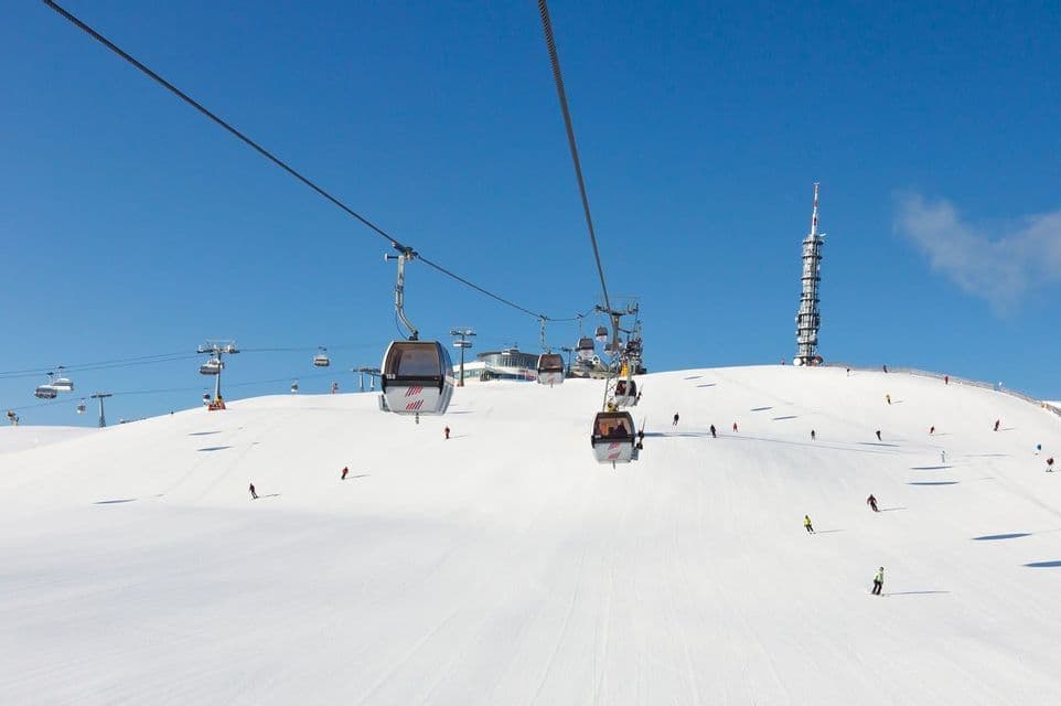 Seilbahnen fahren einen verschneiten Berg hinauf, während Skifahrer unter klarem blauem Himmel die Piste hinuntergleiten.