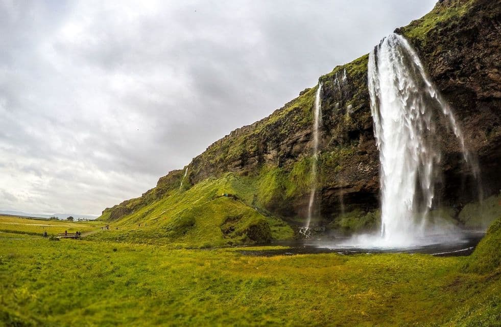 Cascada alta cayendo por un acantilado cubierto de musgo sobre un campo verde exuberante bajo un cielo nublado.