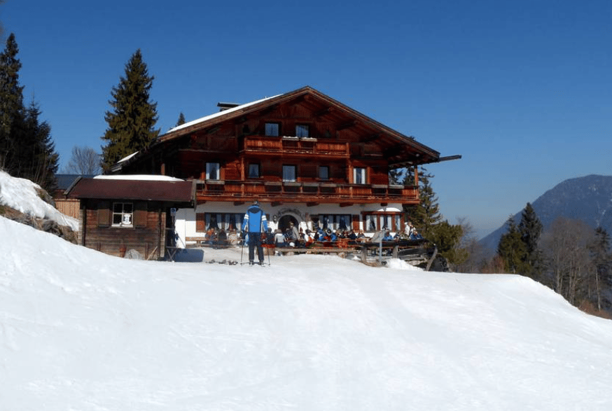 Eine große hölzerne Skihütte an einem verschneiten Berghang mit Menschen, die sich auf der Terrasse unter einem klaren blauen Himmel entspannen.