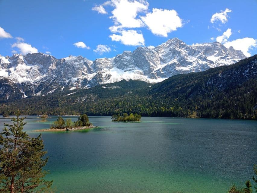 Ein türkisfarbener See mit kleinen, bewaldeten Inseln vor einer großen, schneebedeckten Bergkette unter einem blauen Himmel mit Wolken.