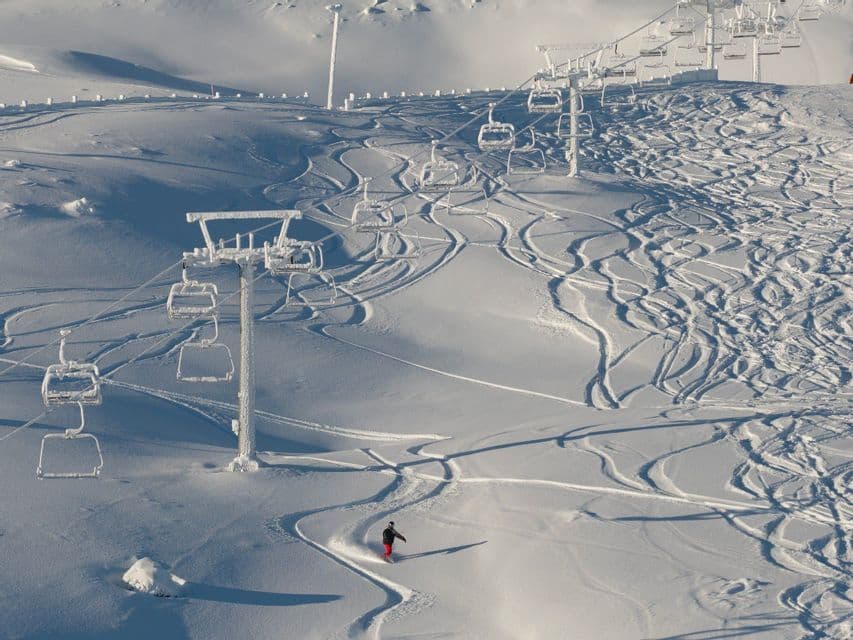 Ein Snowboarder carvt einen verschneiten Hang hinunter, der voller Spuren ist, und gleitet dabei unter leeren, eisbedeckten Sesselliften hindurch.