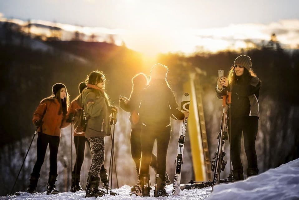 Eine WeRoad-Gruppe in Skiausrüstung steht auf einem verschneiten Hang, während die Sonne untergeht und ein goldenes Gegenlicht erzeugt.