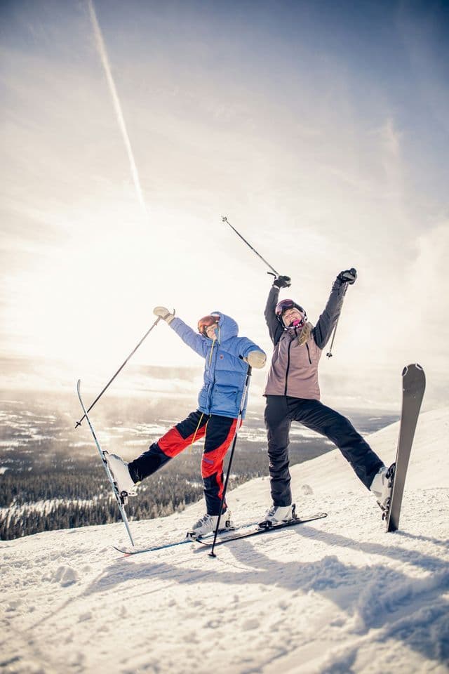Ein Skifahrer und ein Snowboarder einer WeRoad-Gruppenreise posieren mit erhobenen Armen auf einem sonnigen, verschneiten Berggipfel.