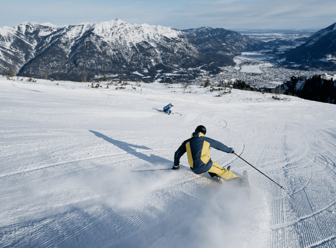 Ein Skifahrer in gelber Jacke carvt eine verschneite Piste hinab, wirbelt Schnee auf, mit Bergen und einem Tal im Hintergrund.