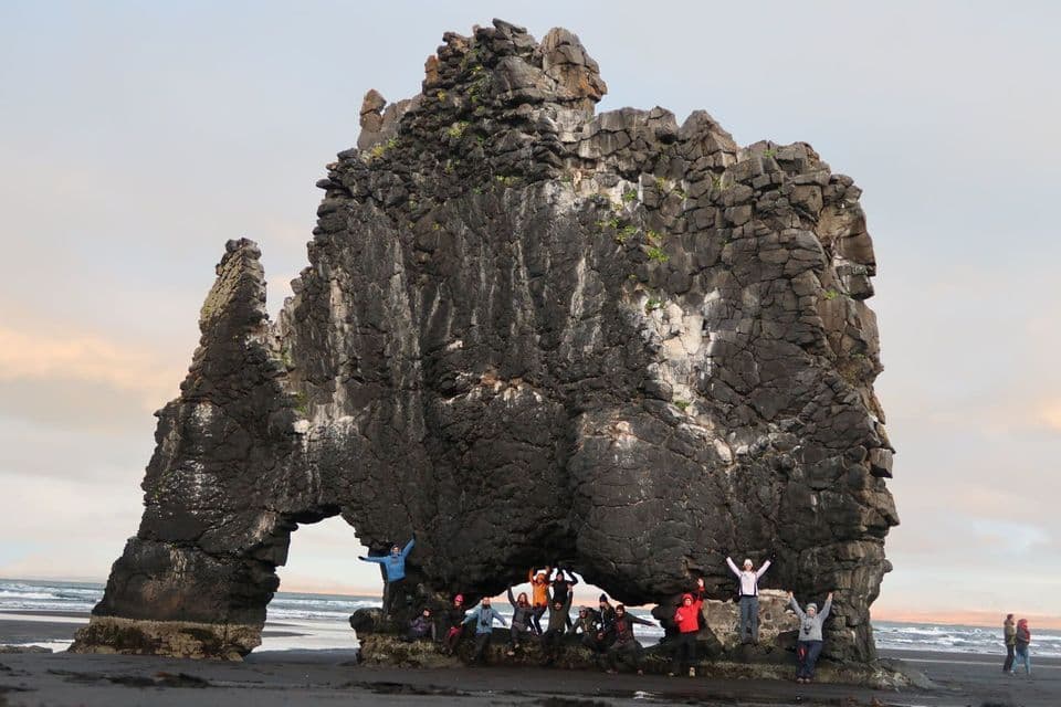 A WeRoad group trip poses for a photo under a large, arched rock formation on a dark sand beach.