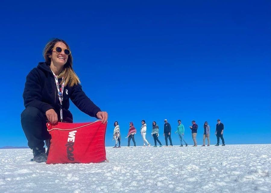 Une femme s'accroupit, tenant un sac WeRoad rouge, sur une vaste étendue de sel blanc, avec le groupe de voyage WeRoad aligné en arrière-plan sous un ciel bleu clair.