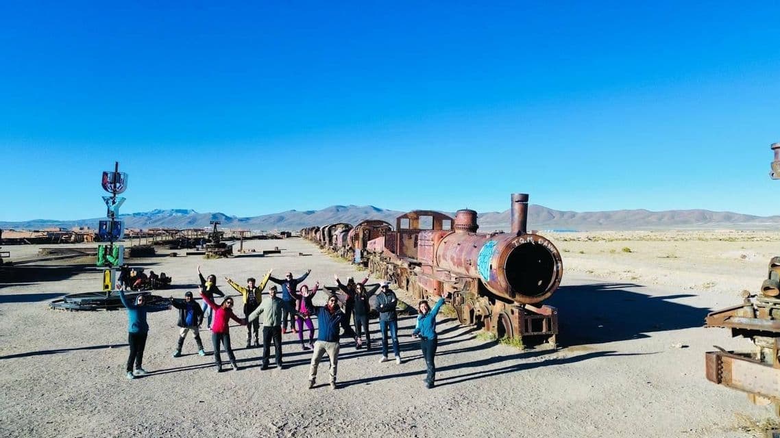A WeRoad group trip poses with arms raised in front of a long, rusty, abandoned train in a desert landscape.