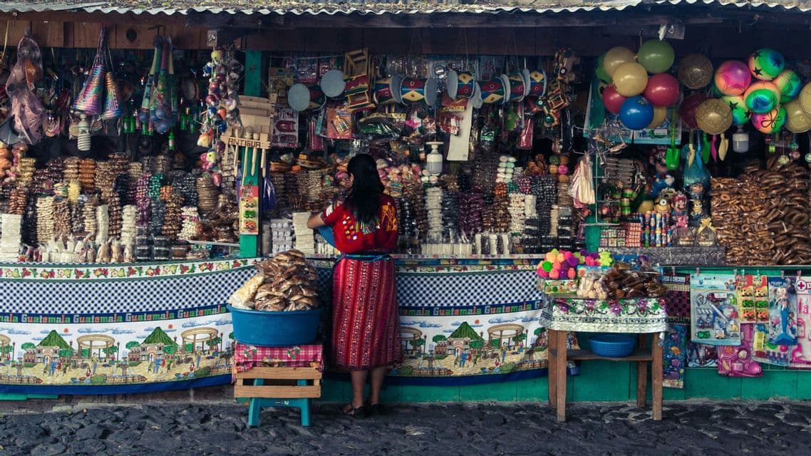 Eine Frau in traditioneller Kleidung steht an einem Marktstand im Freien voller bunter Süßigkeiten, Spielzeug und lokalem Kunsthandwerk.