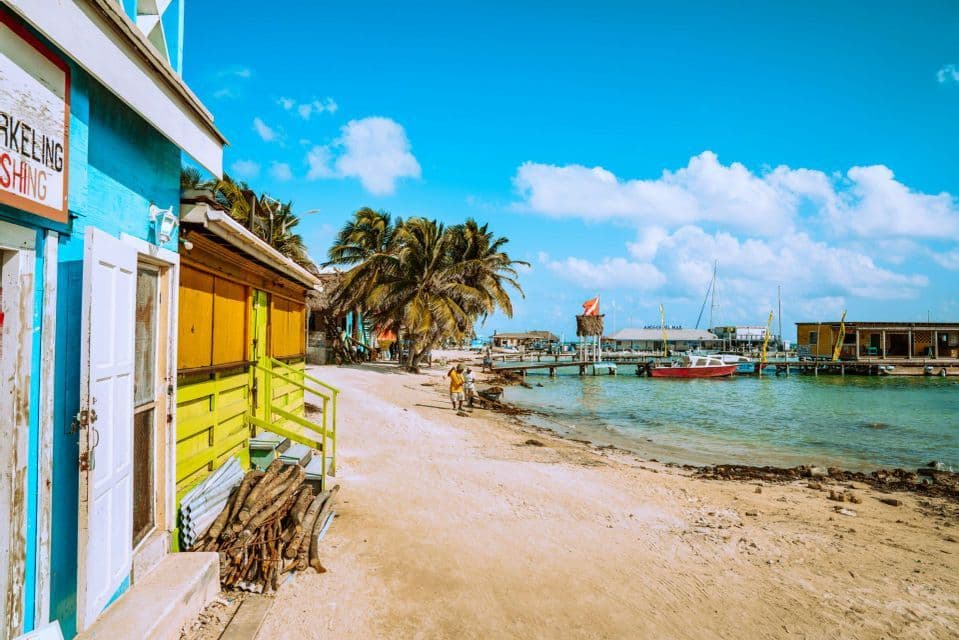 Bunte Holzgebäude säumen einen Sandstrand mit Palmen neben einem türkisfarbenen Ozean mit einem Pier und Booten unter einem sonnigen Himmel.