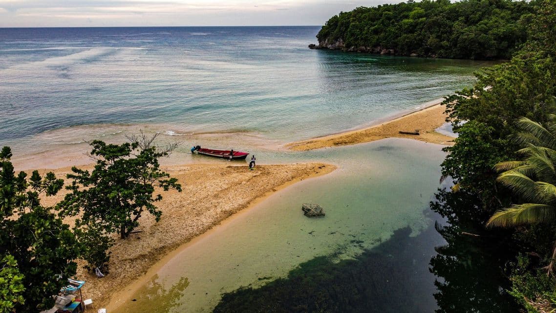 Luftaufnahme einer Sandbank, die eine ruhige Bucht vom Ozean trennt, mit einem kleinen Boot in der Nähe und umgeben von tropischem Wald.